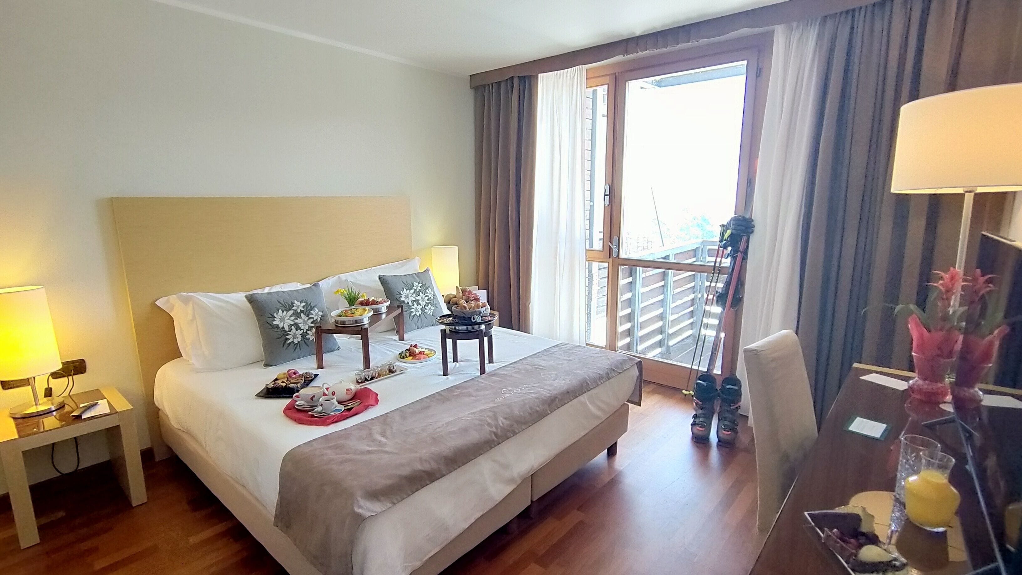 Una habitación de hotel con cama, mesitas de noche, una pequeña mesa con comida, una silla y una ventana con cortinas.