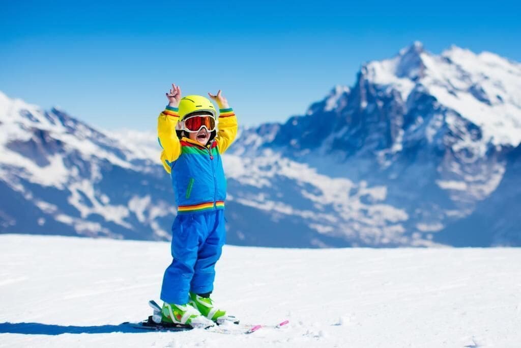 Un enfant fait du ski sur une montagne enneigée, sous un ciel bleu dégagé, avec des sommets couverts de neige en arrière-plan.