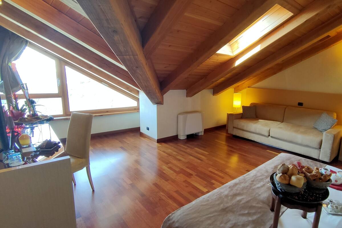 Une pièce chaleureuse de style loft, avec des poutres en bois, un canapé, une table à manger agrémentée d’un panier de fruits et une fenêtre laissant entrer la lumière naturelle.