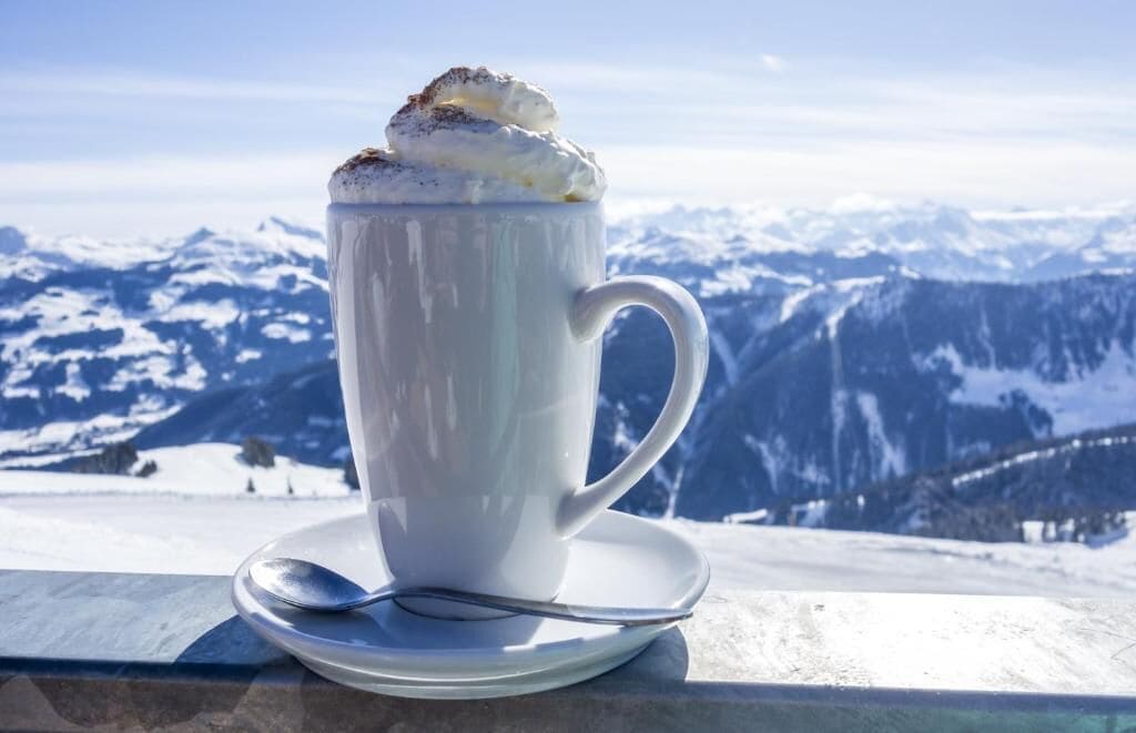 Una tazza bianca con panna montata su un piattino, posizionata su un balcone con vista sulle montagne.