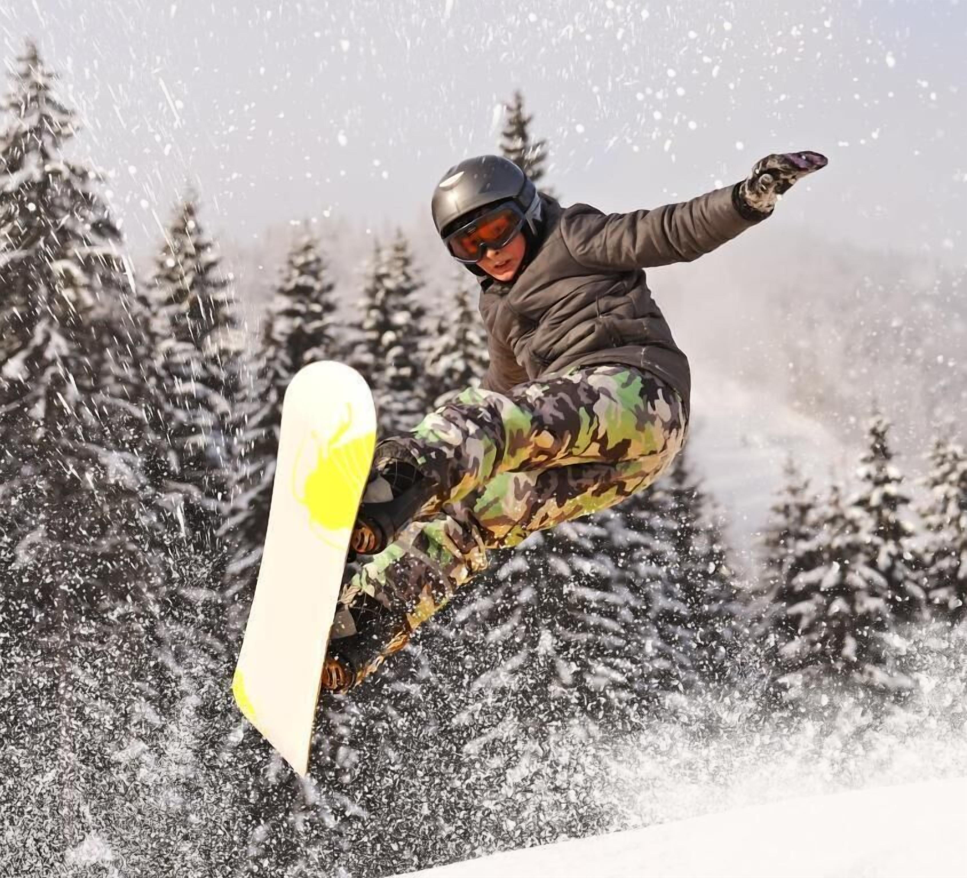 Un snowboarder en pleno vuelo realizando un salto, con árboles al fondo.