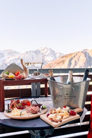Una mesa con una tabla de embutidos, dos copas de vino blanco y un cubo de champán con botellas en un balcón con vistas a la montaña.