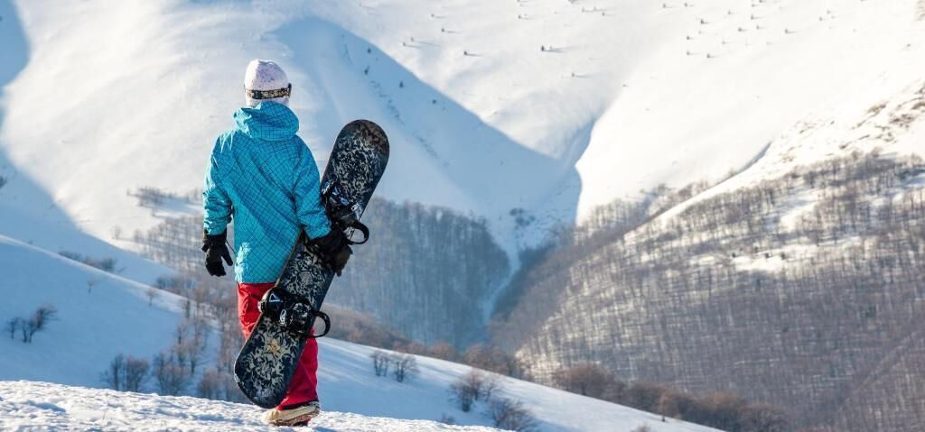 A person standing on a snowy slope holding a snowboard, with a mountainous background.