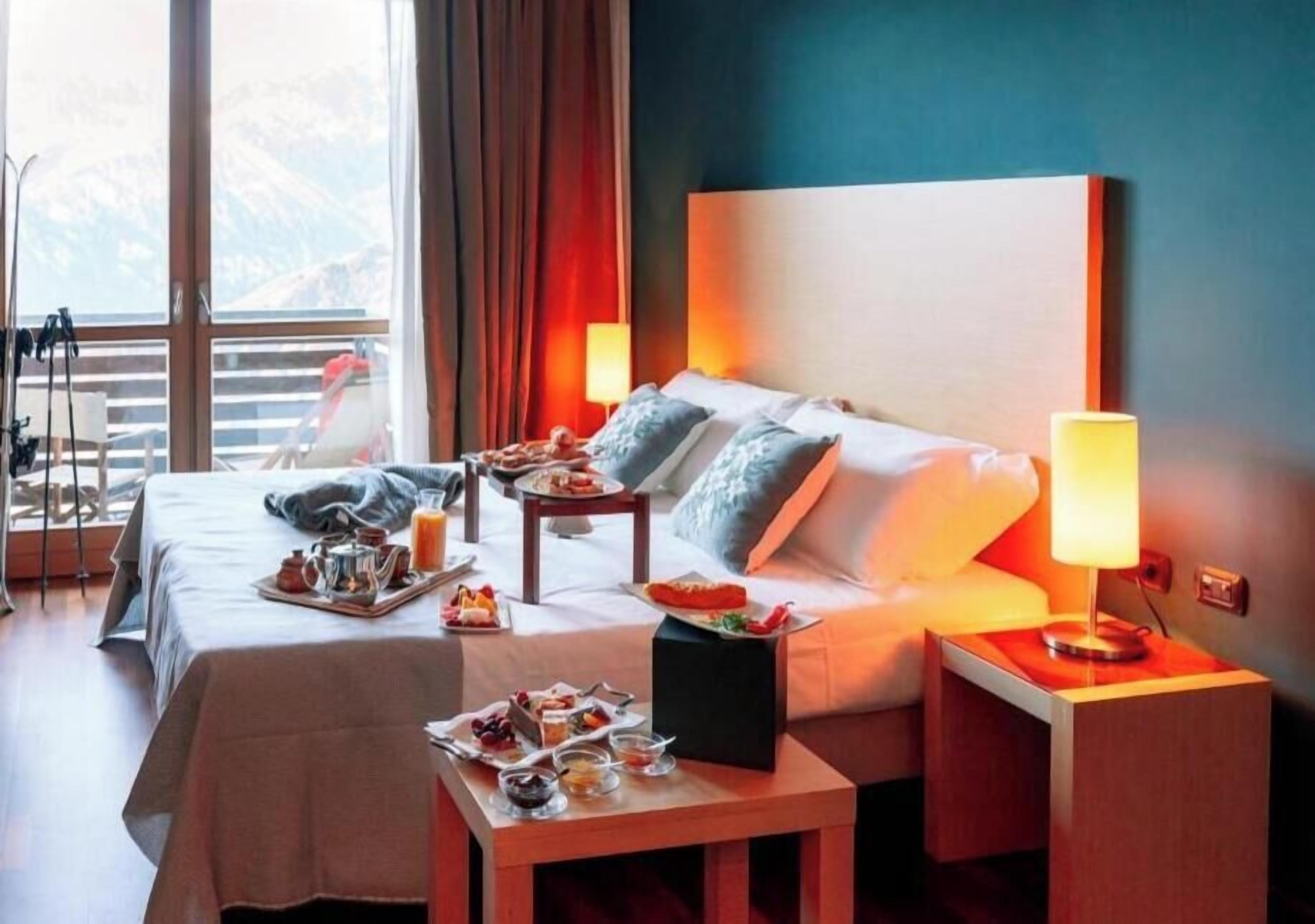 Ein Hotelzimmer mit zwei Betten, einem Esstisch mit Frühstück, einem Nachttisch mit Lampe und Blick auf Berge durch große Fenster.