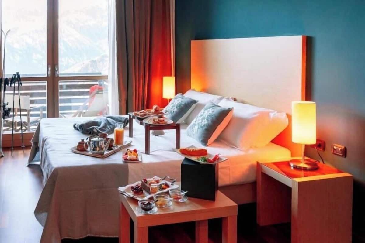 Une chambre d’hôtel avec deux lits, une table à manger où le petit-déjeuner est servi, une table de chevet avec une lampe, et une vue sur les montagnes à travers de grandes fenêtres.