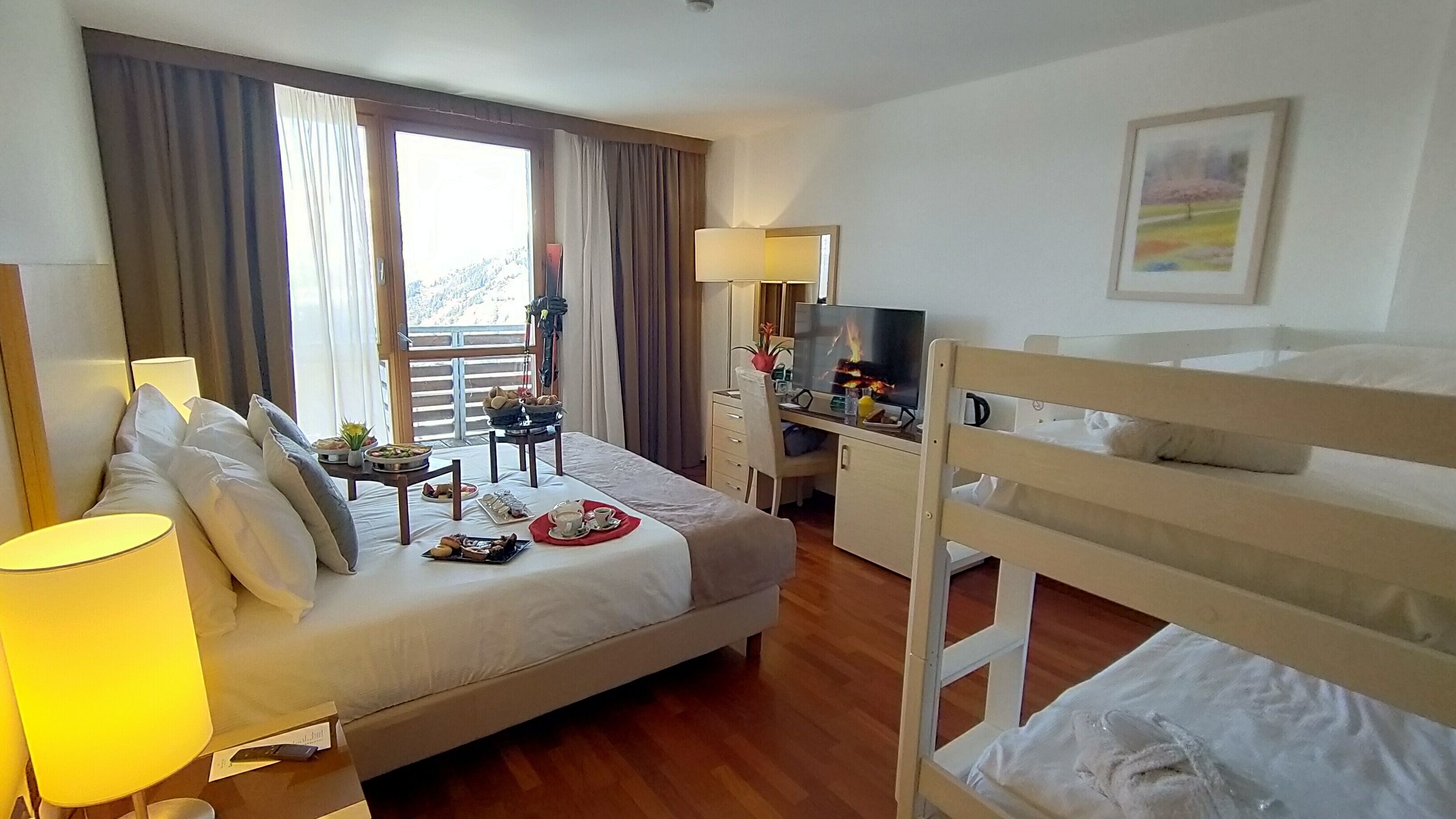 A hotel room with a bed, a desk with a TV, a balcony with curtains, and a painting on the wall.