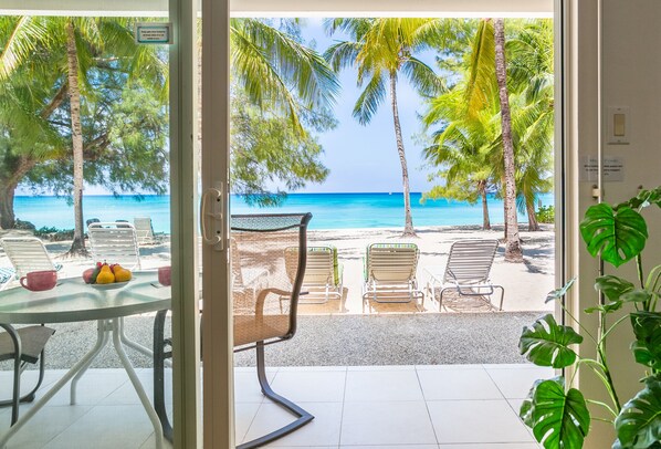 A view from a room with a glass door to a beach, featuring a table with fruits, chairs, and a view of the ocean and palm trees.
