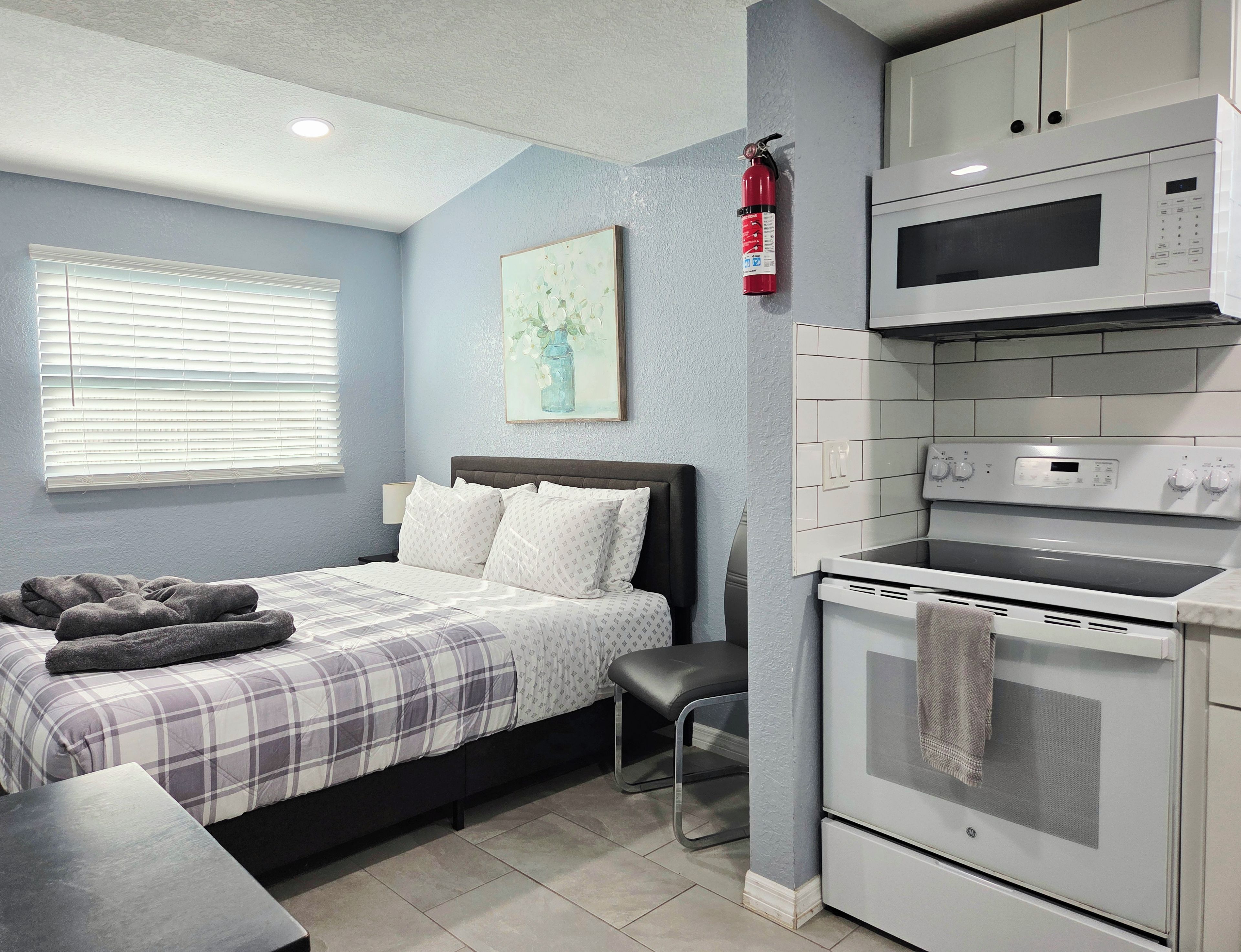 Un dormitorio con cama, mesita de noche y un pequeño taburete. Hay una kitchenette con microondas y horno.