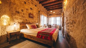 Quarto com paredes de pedra, cama de madeira, mesa de cabeceira, abajur e janela com cortinas.