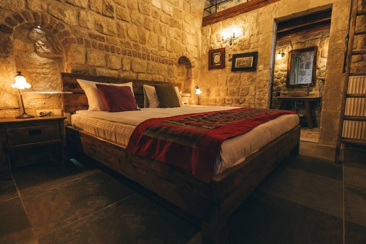 A cozy bedroom with a stone wall, a wooden bed, and a nightstand with a lamp.