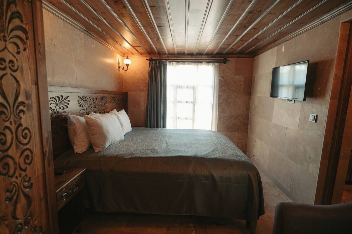 A room with a wooden ceiling, a bed with a headboard, a flat-screen TV mounted on the wall, and a window with curtains.