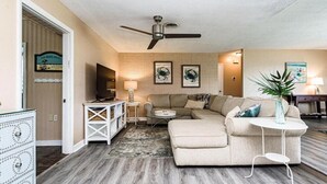 A living room with a beige sofa, a coffee table, a TV, and a ceiling fan. - Private 4 bedroom Pool Home w 2 car garage, Walk to the Beach, Dog Friendly (Ormond Beach)