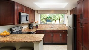 A compact kitchen with dark wood cabinets, a granite countertop, a built-in microwave, a stove, a sink, and a refrigerator. There are two stools and a window with a view of greenery outside. - Sheraton Vistana Resort in Orlando, Florida (includes Christmas weekend) (Orlando)