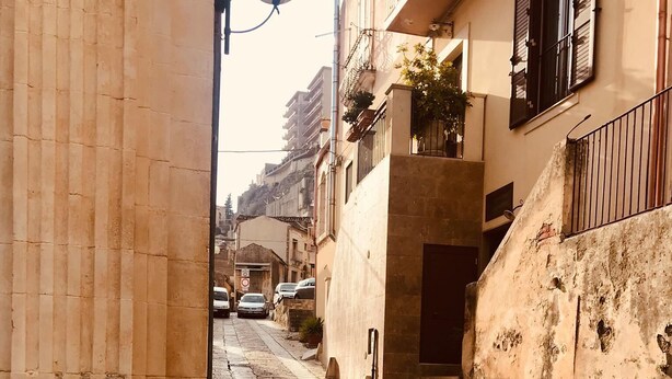 A narrow street with stone pavement, buildings on either side, and a metal gate in the foreground. - Vacation Home PETRA (Modica)