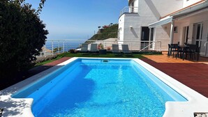 En pool med udsigt til havet, en patio med stole og en bygning med balkon.