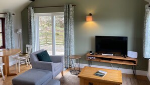 Un salón con un sofá gris, una mesa de centro de madera, un televisor de pantalla plana sobre un mueble de madera y un amplio ventanal con cortinas.