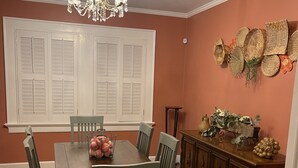 Une salle à manger avec une table et des chaises en bois, un lustre, une fenêtre avec des volets et un mur décoré de paniers tressés.