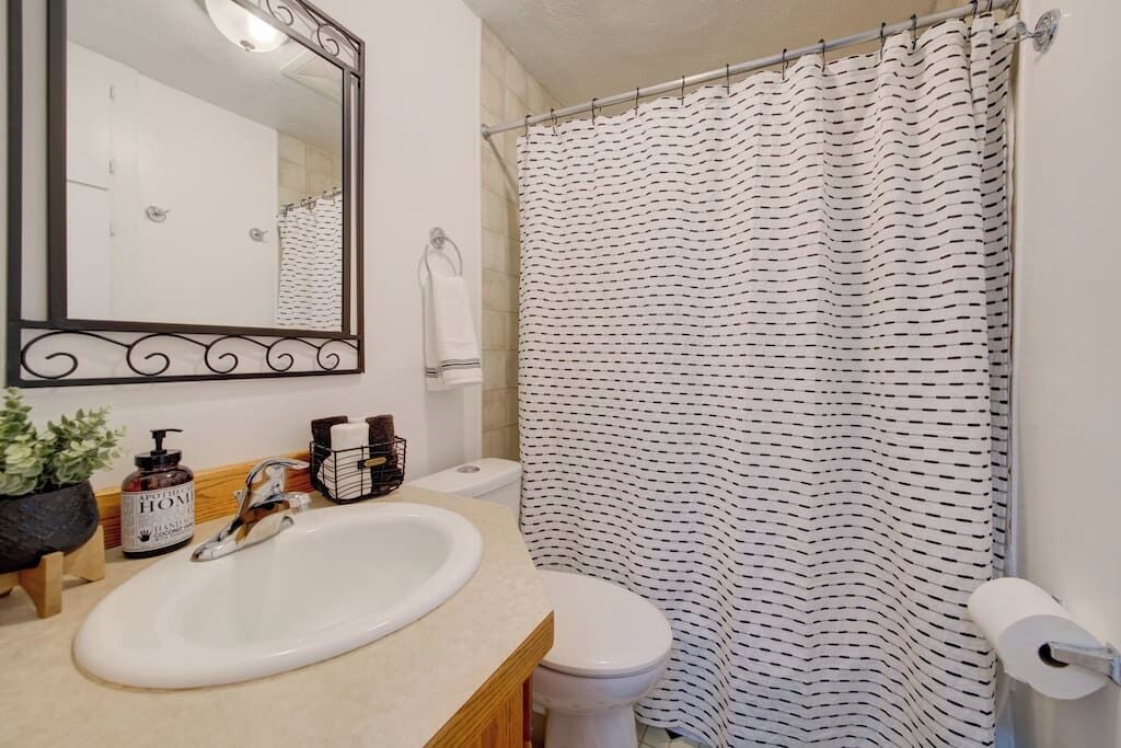 A bathroom with a white sink, a toilet, a shower curtain with a black pattern, a mirror with a decorative frame, and a small potted plant.