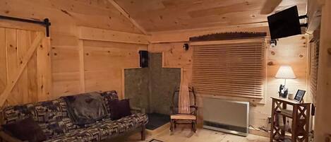Ein gemütliches Blockhaus-Interieur mit Holzbalkendecke, Deckenventilator, einem gemusterten Teppich, einer Couch, einem Sessel, einem kleinen Tisch mit Lampe und einem an der Wand montierten Fernseher.