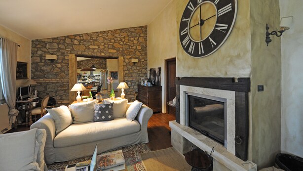 A living room with a stone wall, a fireplace, a large wall clock, and a sofa. - Luxury Country House in Tuscany (Semproniano)