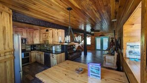 Un interior de cabaña espacioso con paredes y suelos de madera, una zona de cocina, una mesa de comedor y una sala de estar con chimenea.