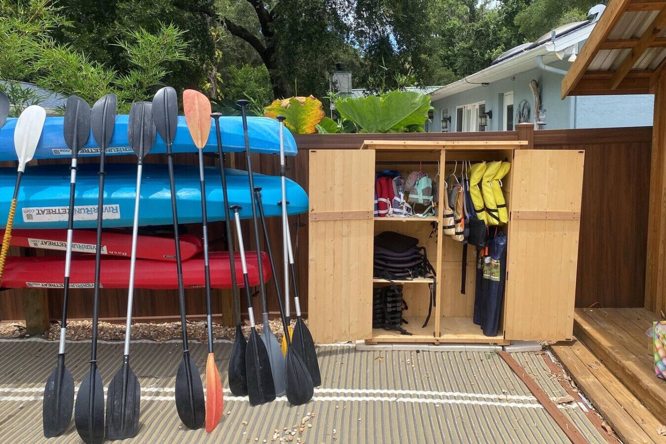 Un espacio exterior cubierto con un cobertizo de madera que contiene kayaks y paletas, un pequeño cuarto de almacenamiento con bolsas y equipo, y un edificio con techo inclinado al fondo.