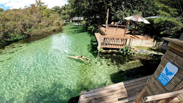 Cuerpo de agua cristalina con una persona nadando, rodeado de estructuras de madera y vegetación densa.