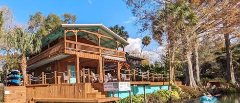 A wooden cabin with a balcony, situated on stilts over water, surrounded by trees.
