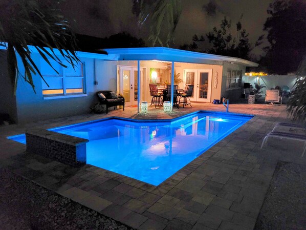 Une piscine avec une terrasse carrelée, un patio couvert aménagé avec du mobilier d’extérieur, et une maison dont on peut voir la porte et les fenêtres.