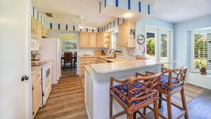 A kitchen with wooden cabinets, a central island with bar stools, and a view of the outdoors through a window. - Cool Breeze. A comfortable house with a tropical backyard waterfront view.  (Naples)