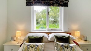 Ein Schlafzimmer mit einem Bett, zwei Nachttischen, Lampen, einem Fenster mit Blick auf Grünflächen und einem gemusterten Kissen.