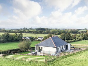 A house with a large deck, surrounded by a wooden fence, set in a rural landscape with open fields and distant hills.