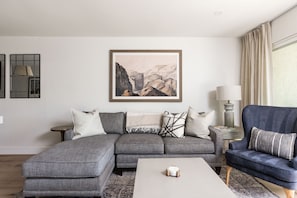 A modern living room with a grey sectional sofa, a blue armchair, a coffee table, and a large landscape painting on the wall.