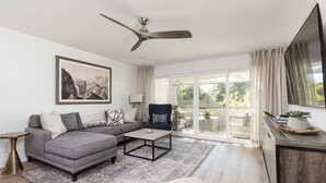 A modern living room with a grey sectional sofa, a coffee table, and a ceiling fan. There is a large window with curtains, a TV, and a plant on a wooden table. - Ultra-Clean, Designer Condo in Downtown Scottsdale (Scottsdale)