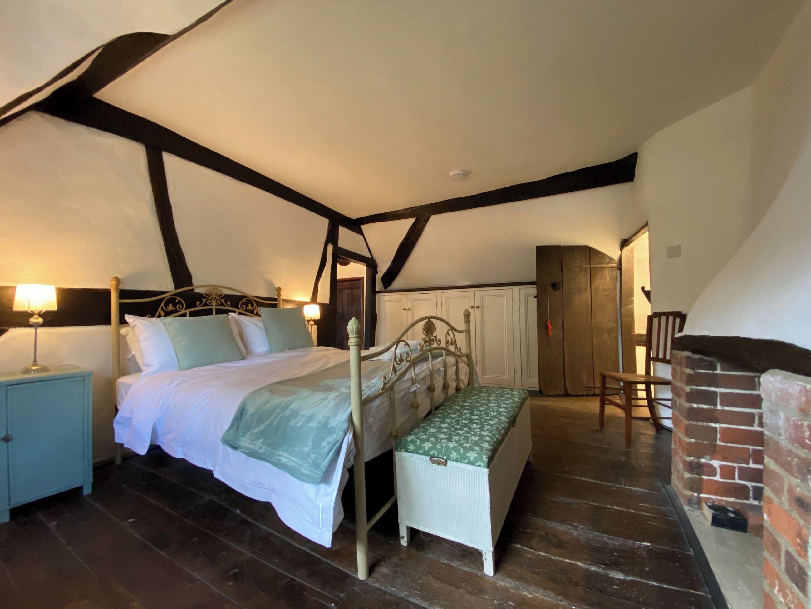 Quarto com uma cama de dossel, um banco de madeira, uma mesa de cabeceira e uma lareira de tijolos.
