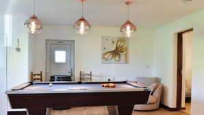 A pool table with three hanging lights, a wooden floor, a door with a window, and a painting on the wall.