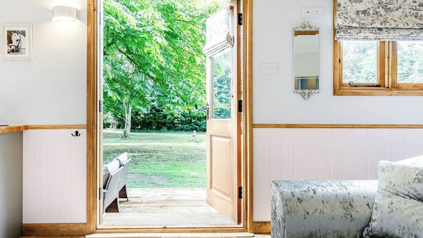 A room with a wooden door leading to a garden, a white cushioned bench, and a patterned sofa. - A lovely shepherd’s hut, set quietly in a paddock with shade from mature trees. (Alkborough, near Scunthorpe)