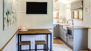 A modern kitchen with a built-in oven, microwave, and sink. There is a wooden table with two stools and a flat-screen TV mounted on the wall. - A lovely shepherd’s hut, set quietly in a paddock with shade from mature trees. (Alkborough, near Scunthorpe)