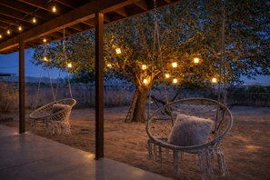 A covered outdoor area with two swing chairs and hanging lights. - Relax in a peaceful Yucca Valley desert retreat just minutes from Joshua Tree. Enjoy a private yard, optional hot tub under the stars, fire pit, and stylish, cozy interiors—perfect for stargazing, unwinding, and exploring the High Desert. (Yucca Valley)