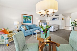 A modern hotel room with a blue sofa, a wooden coffee table, a large bed, and a painting of a beach scene.
