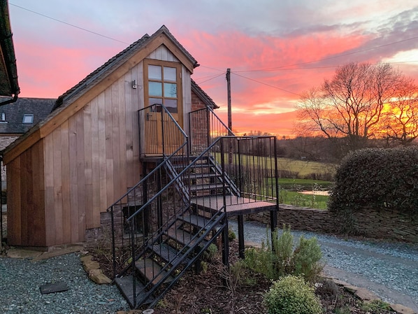 A wooden cabin with a staircase leading to a balcony, surrounded by a gravel path and greenery.
