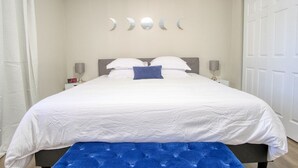 A neatly made bed with white bedding and a blue accent pillow, a bedside table with a lamp, and a wall-mounted crescent moon decoration. - Modern-Sunroom-15min to DwnTwn-Grps-Kids-Firepit (Indianapolis)