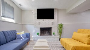 A living room with a white brick fireplace, a mounted TV, a blue sofa, a yellow sofa, and a patterned rug. - Modern-Sunroom-15min to DwnTwn-Grps-Kids-Firepit (Indianapolis)