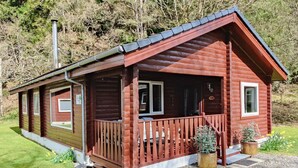 Une cabane en bois dotée d’un porche, d’une cheminée et d’un petit jardin agrémenté de plantes en pot.