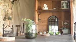 A rustic room with a wooden table, a glass jar with plants, a wooden chair, a shelf with decorative items, and a window with a view of greenery. - Charming gîte with garden and fireplace (Neuilly-en-Vexin)