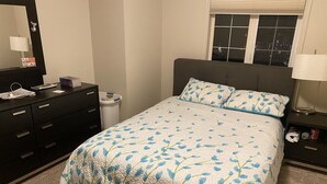 Ein Schlafzimmer mit Bett, Kommode, Nachttisch, Fenster und Spiegel.