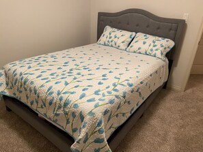 Un lit avec une couvre-lit à motifs floraux et des coussins assortis, une tête de lit capitonnée et un sol recouvert d’une moquette.