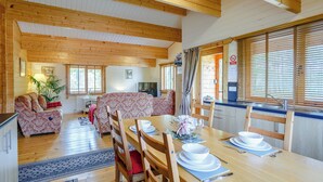 A wooden interior with a dining table set for four, a sofa, a TV, and a window with blinds.