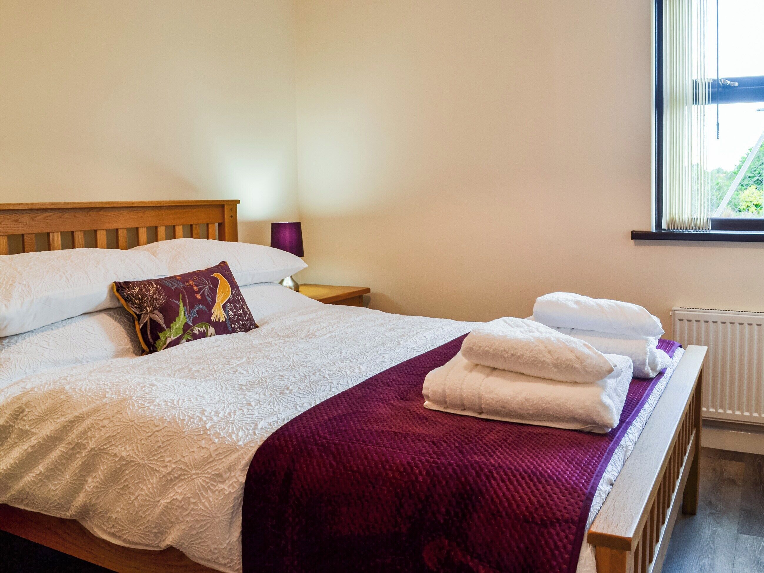 A neatly made bed with white linens and a purple throw blanket, a wooden headboard, a bedside table with a lamp, and a window with blinds.