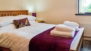 A neatly made bed with white linens and a purple throw blanket, a wooden headboard, a bedside table with a lamp, and a window with blinds. - Keepers Retreat, The Annex (Roughton)